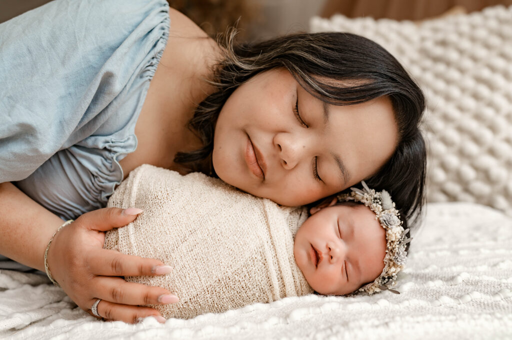 Mother in soft blue dress holding newborn lying on a soft bed, with their cheeks touching and eyes closed in natural light during a fully styled studio newborn session in Annapolis, Maryland.