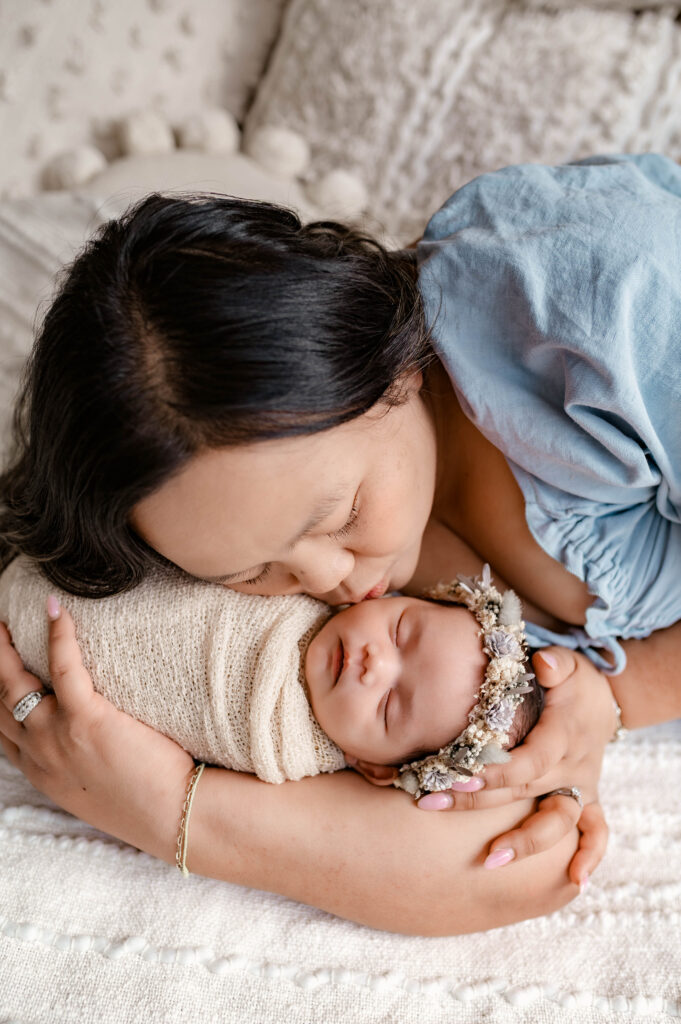 Mother in soft blue dress holding newborn lying on a soft bed, gently kissing her cheek in natural light during a fully styled studio newborn session in Annapolis, Maryland.