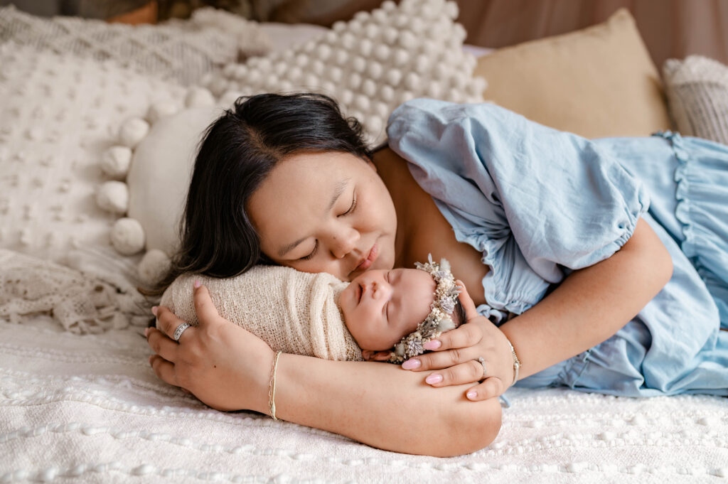 Mother in soft blue dress holding newborn lying on a soft bed in natural light during a fully styled studio newborn session in Annapolis, Maryland.
