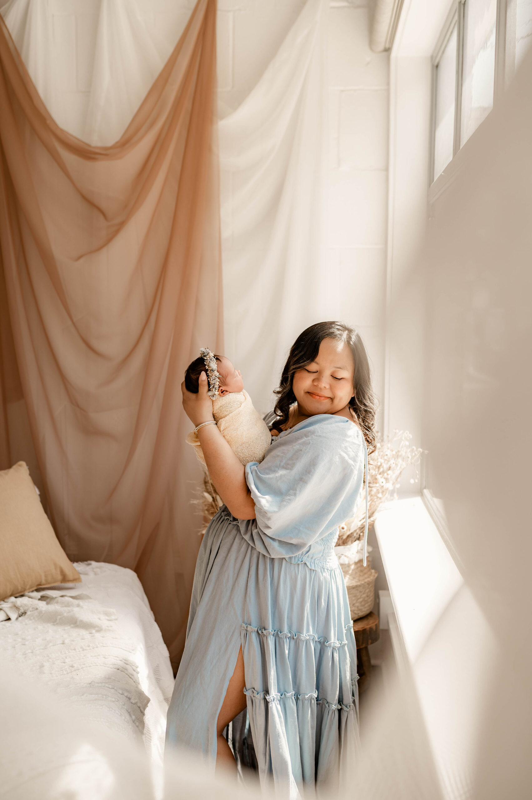 Mother in soft blue dress holding newborn in natural light during a fully styled studio newborn session in Annapolis, Maryland.
