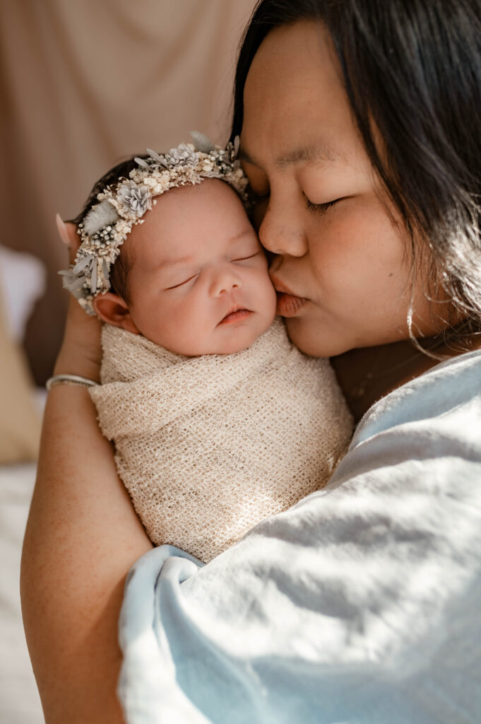 Mother in soft blue dress holding newborn standing by a window, gently kissing her cheek in natural light during a fully styled studio newborn session in Annapolis, Maryland.