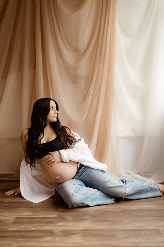 woman sits on floor of photo studio with her hand on her pregnant belly, legs off to the side and she looks towards the light