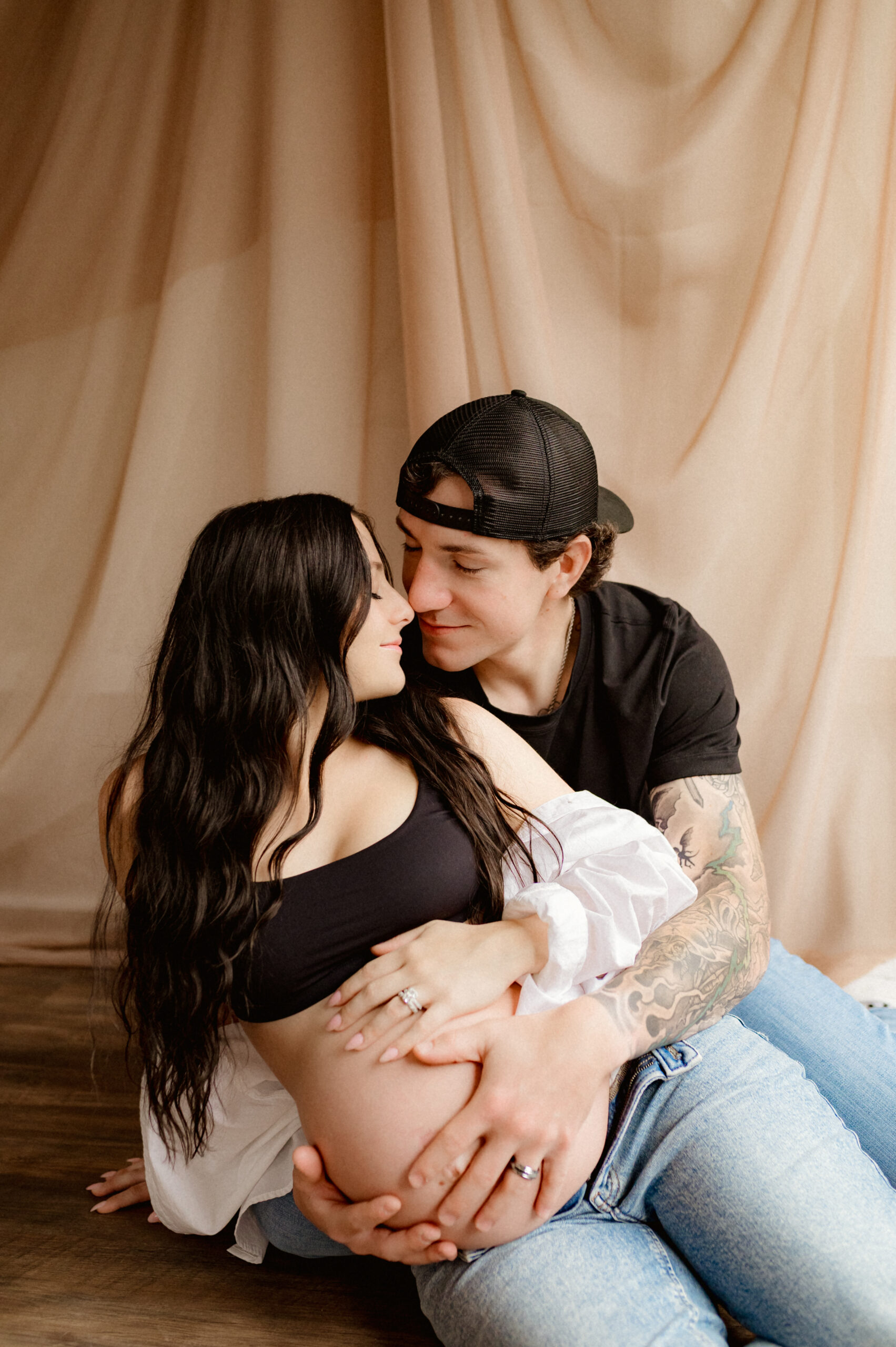 Man sitting behind his partner on the floor, arms gently wrapped around her pregnant belly, noses touching as they look into each other’s eyes.