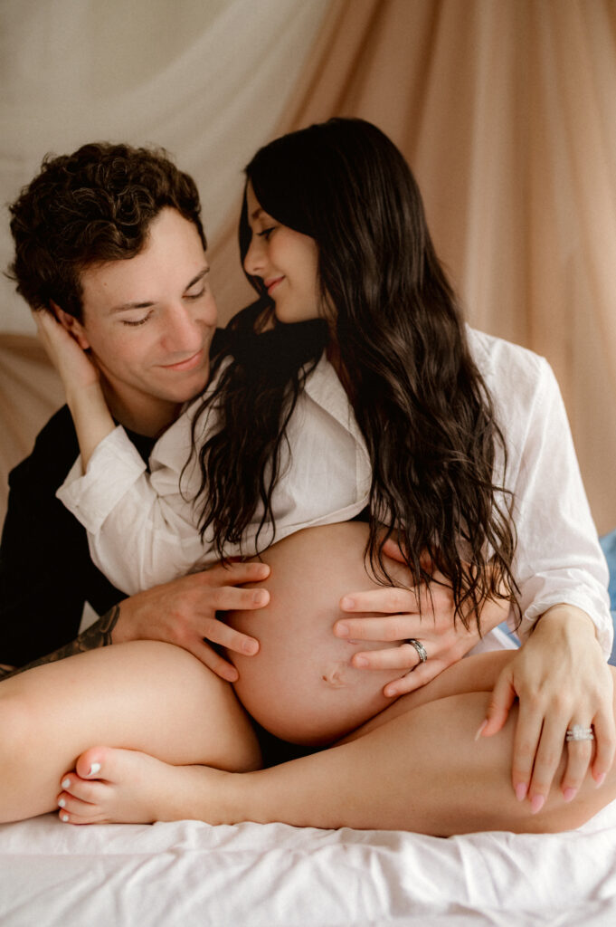 Man sitting behind his partner on a bed, arms gently wrapped around her pregnant belly, her hand is on his face