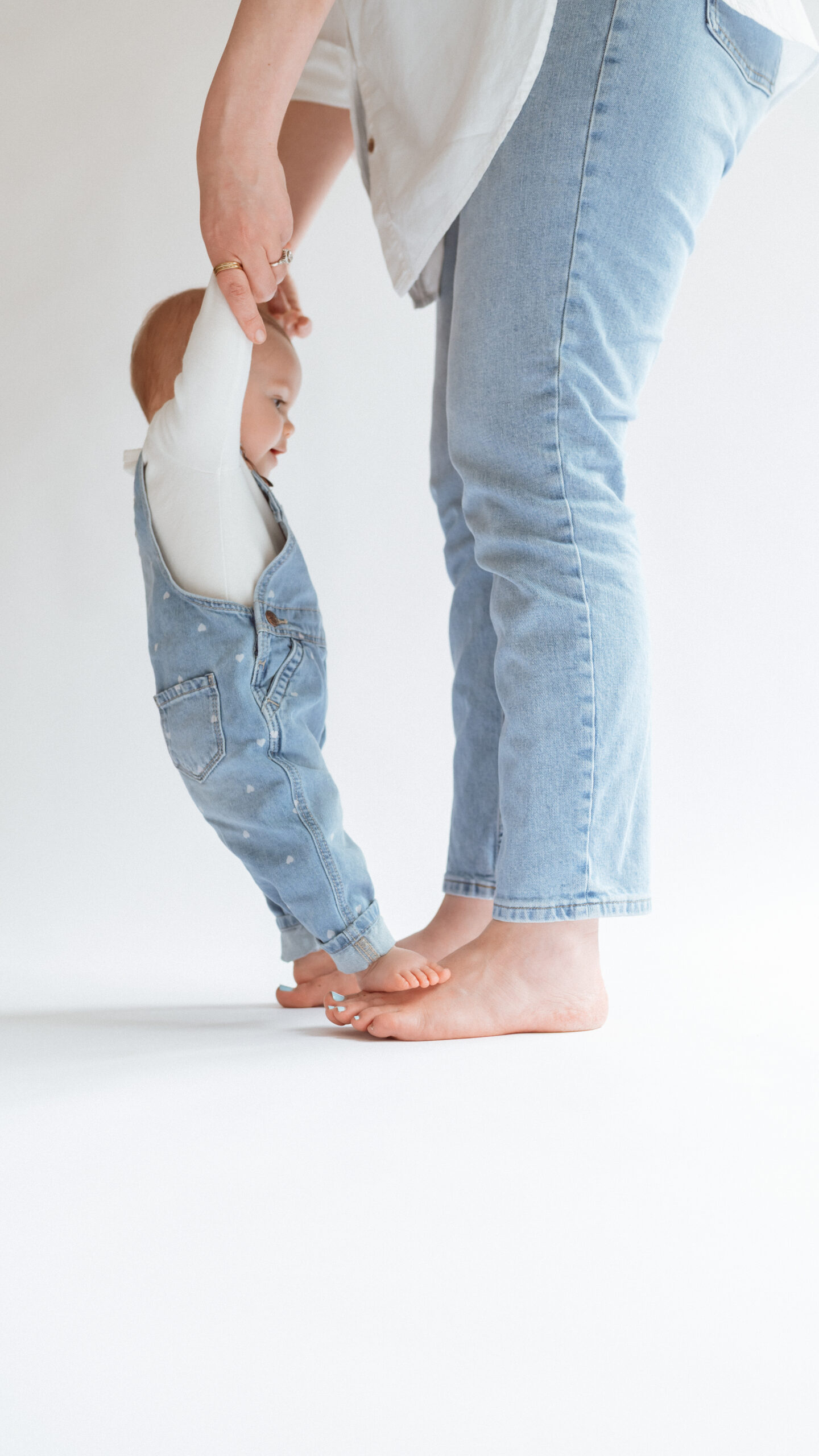 Barefoot mother holding baby’s hand while toddler stands on her feet, both in light denim – modern minimal motherhood photography in Annapolis studio.