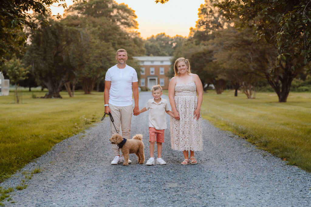 family standing together with their dog on a tree-lined gravel lane in front of farmhouse