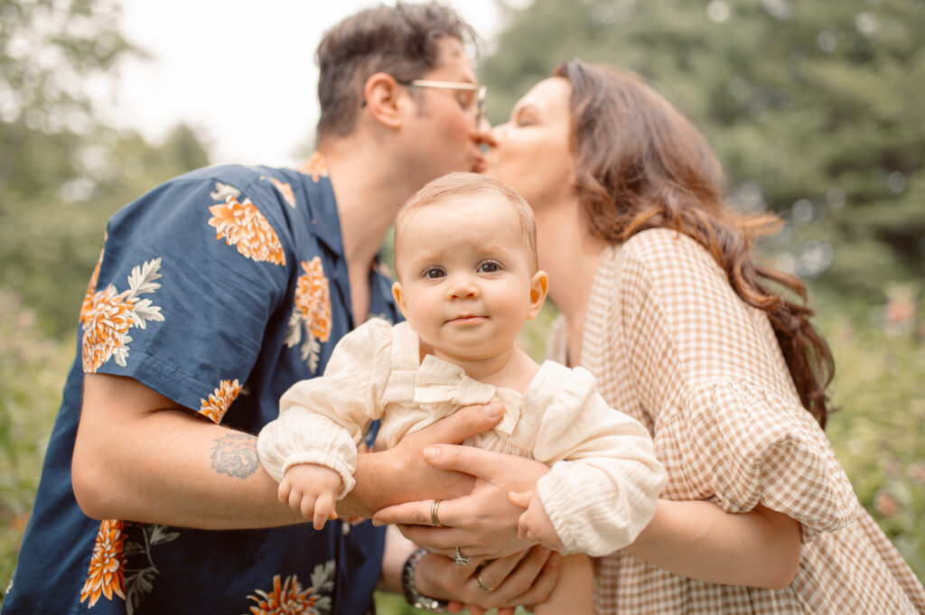 parents share a kiss while they raise their baby girl toward the camera and she smirks right at the lens