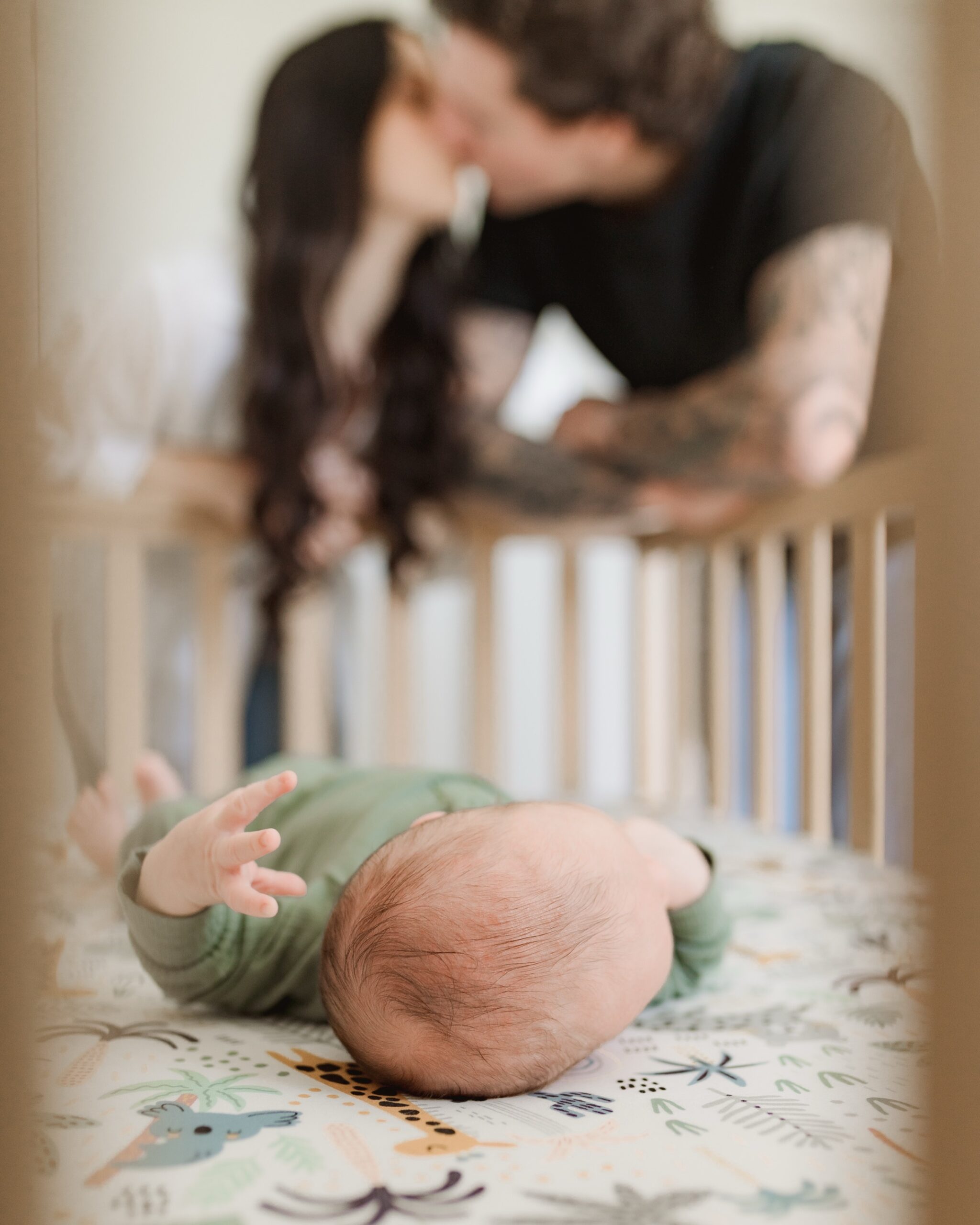 lose-up of newborn baby’s head through crib rails with blurred parents in the background lovingly looking down, captured during an in-home lifestyle session.