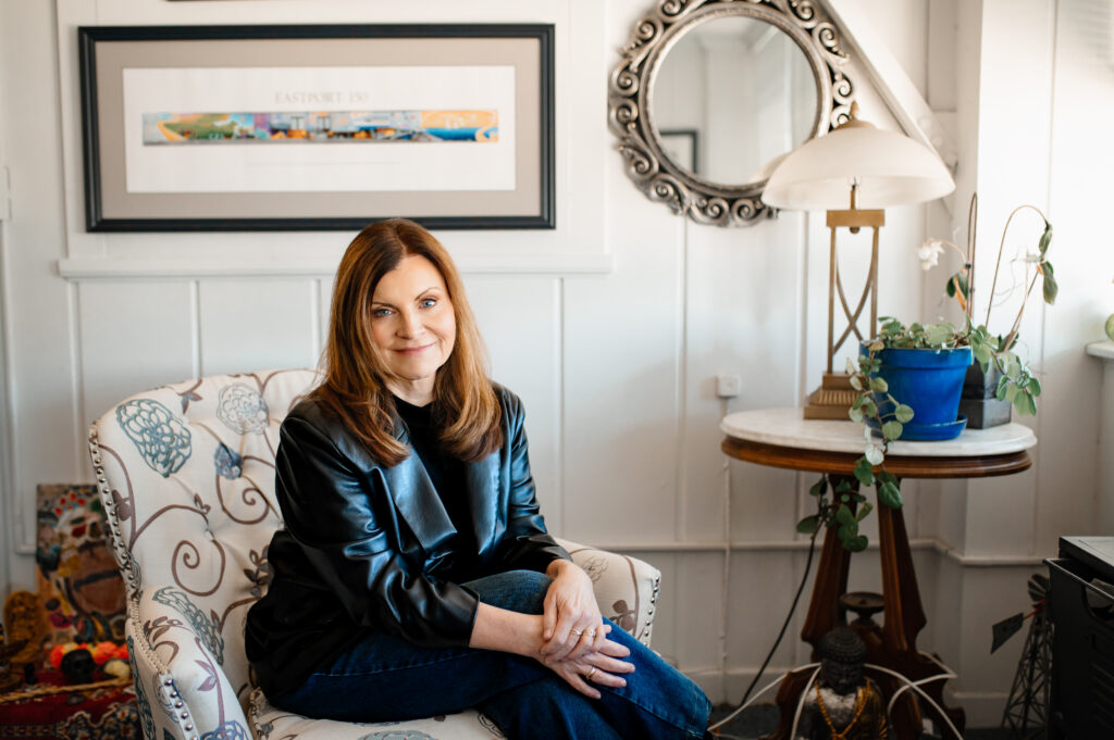 Entrepreneur sitting in her unique office space on a floral chair, smiling directly at the camera. naturally lit image with window to the left.
