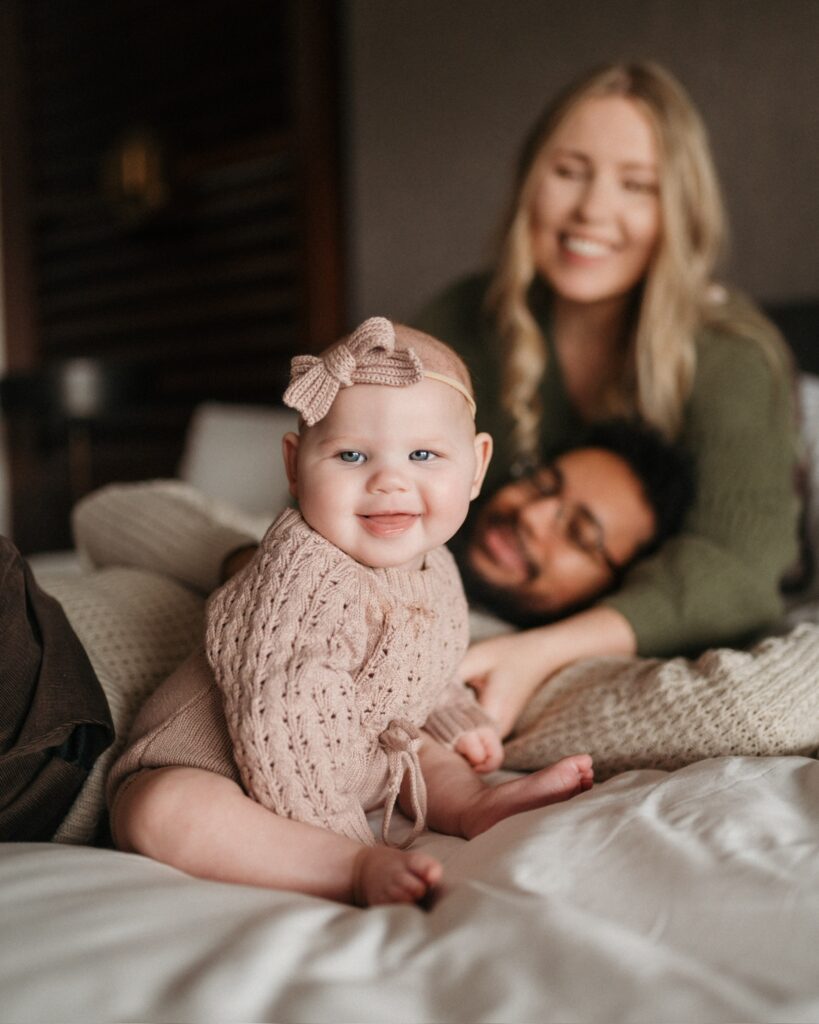 smiling baby girl sitting unassisted on a bed with her parents smiling at her in the background of the image