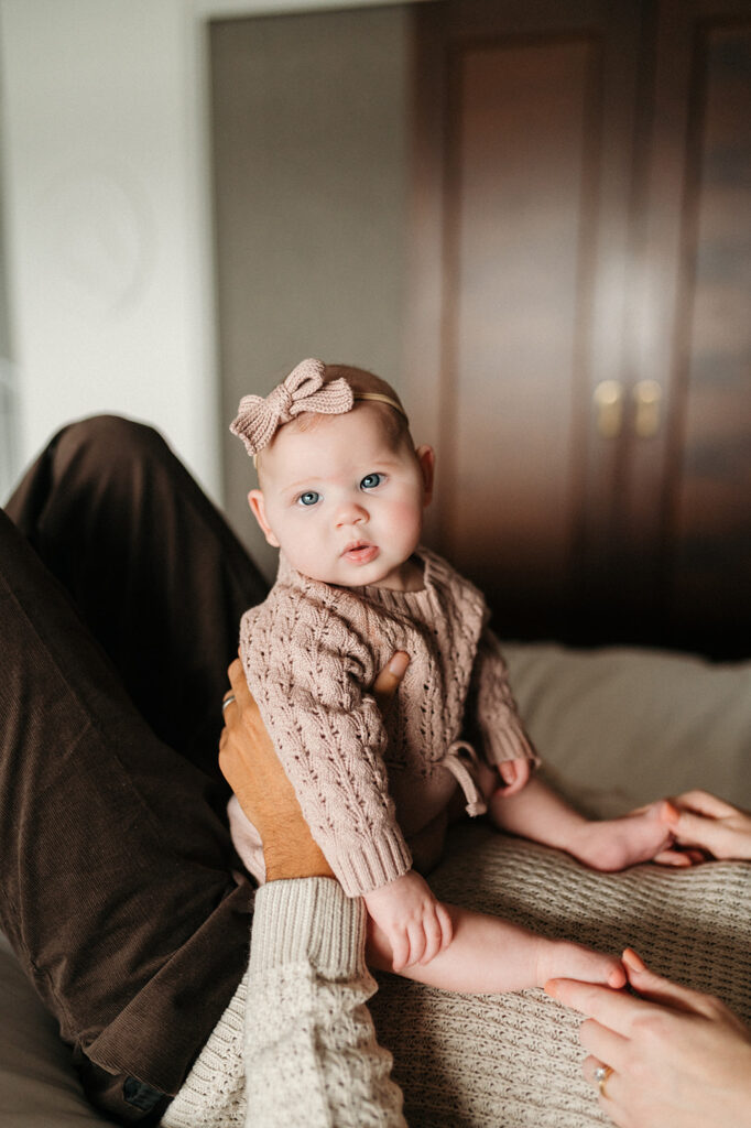 baby sitting on dad's stomach, who is lying down on the bed and his face is out of the frame. she gazes at the camera with a curious look