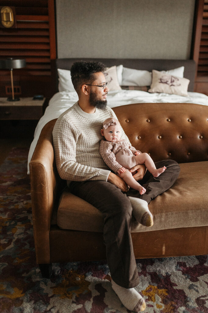 dad sits on couch with one leg tucked under his other leg, baby in lap. she is looking to camera right (mom is next to photographer). dad is gazing out the window