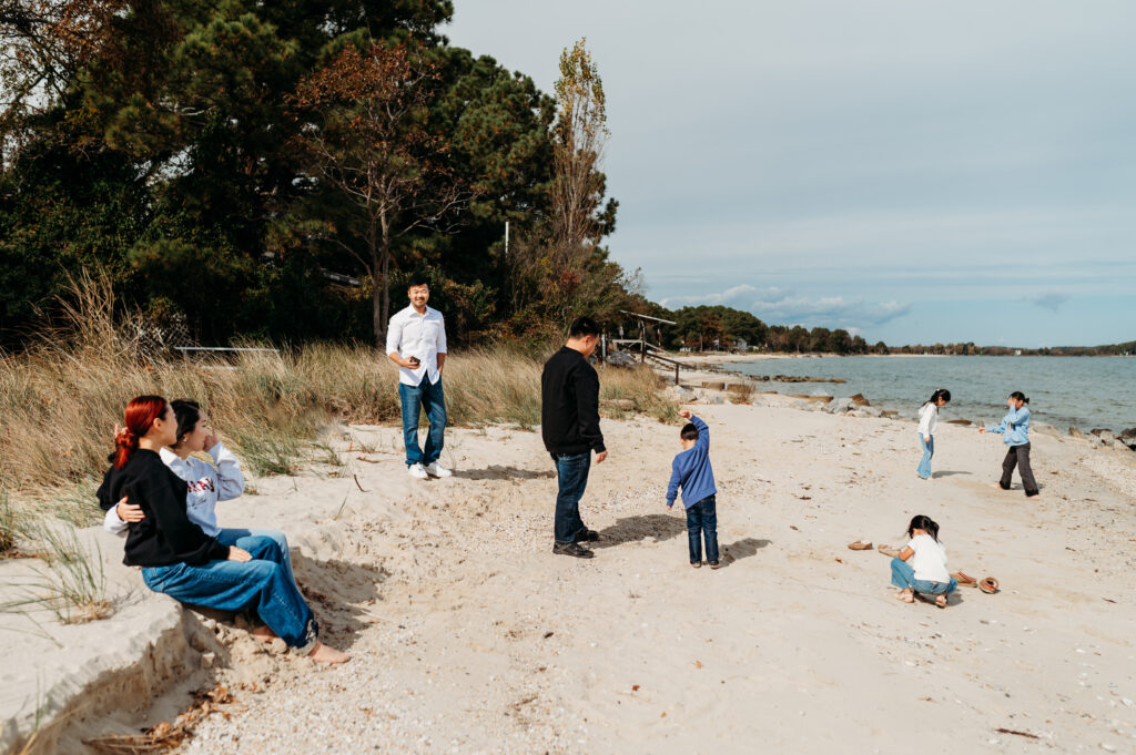 candid image of the entire group of people at this session standing on the beach. On the left are two women, one is wiping tears because she is emotional looking at her family and grateful to be there with her best friend. the fathers are toward the center, the young kids are playing in the sand.