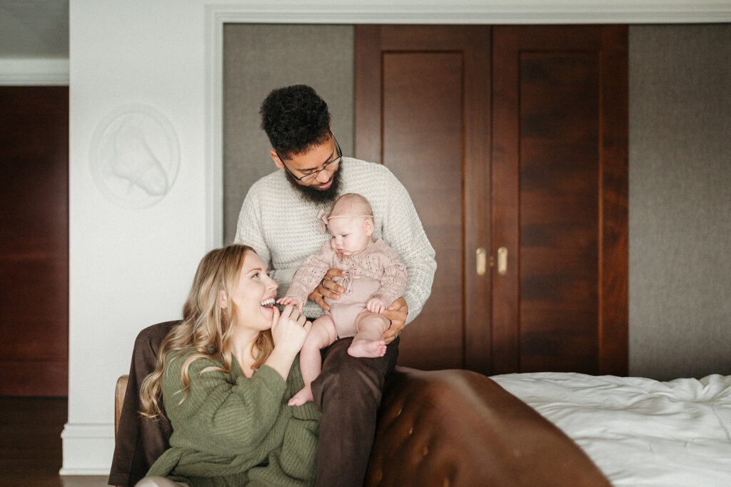 parents snuggling their baby while sitting on a couch in a neutrally styled bedroom setting