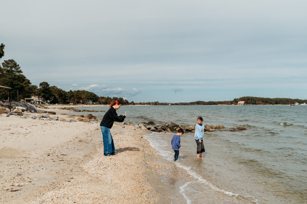 candid moment of a mom taking a photo of her two kids with her phone who are playing in the sand/chesapeake bay, midday and overcast