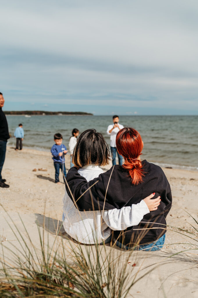 image taken from behind two women who are seated on a deck, holding each other while they look out towards their families, who are playing in the sand