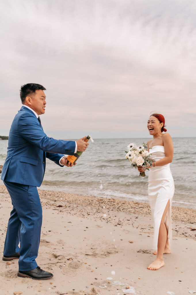 a couple stands on the beach facing each other, while the man in the photo, who is wearing a navy blue suit, is popping a sparkling wine bottle, both are laughing and the bubbly is spilling out. she holds a bouquet and is wearing a strapless white dress