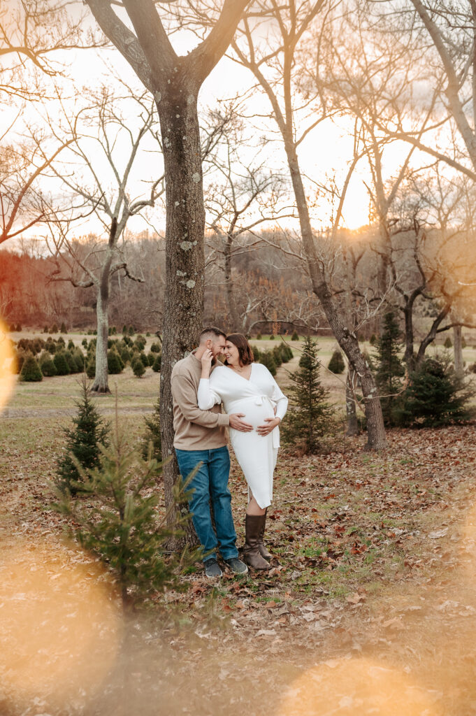 A male and female leaning against a tree embracing while at a farm that has christmas trees all around