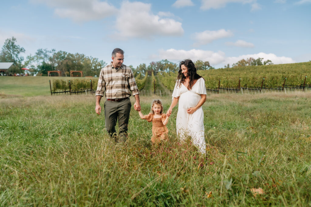 family of 3 walking in a vineyard with a bright blue sky. mom is holding her expectant belly.