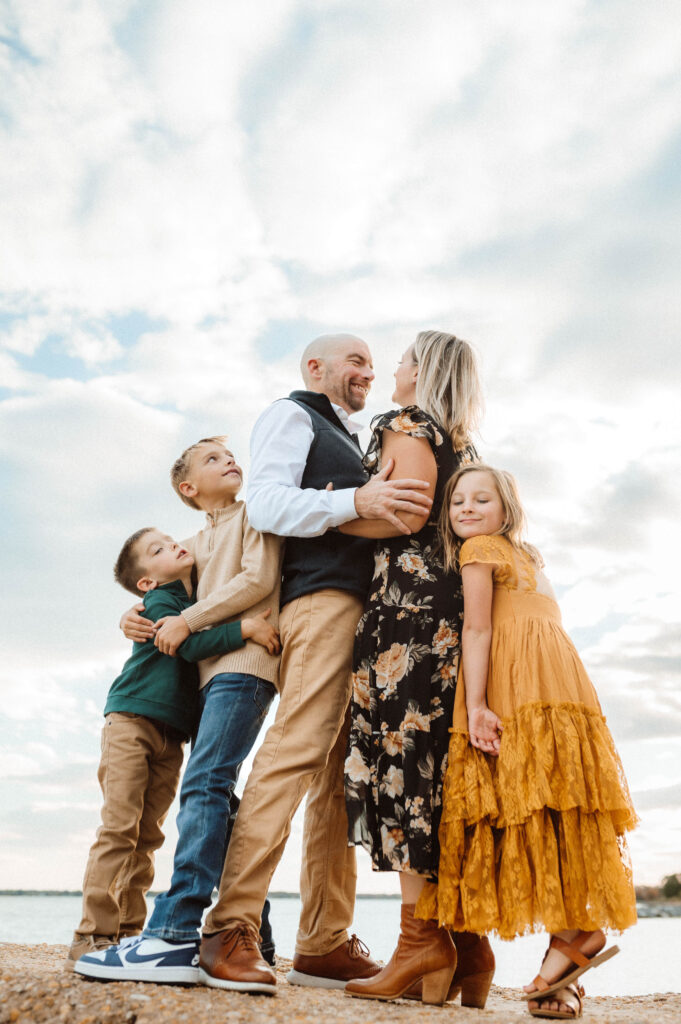 a low perspective image of a family of 5 hugging each other tight on top of a jetty with the clouds framing them