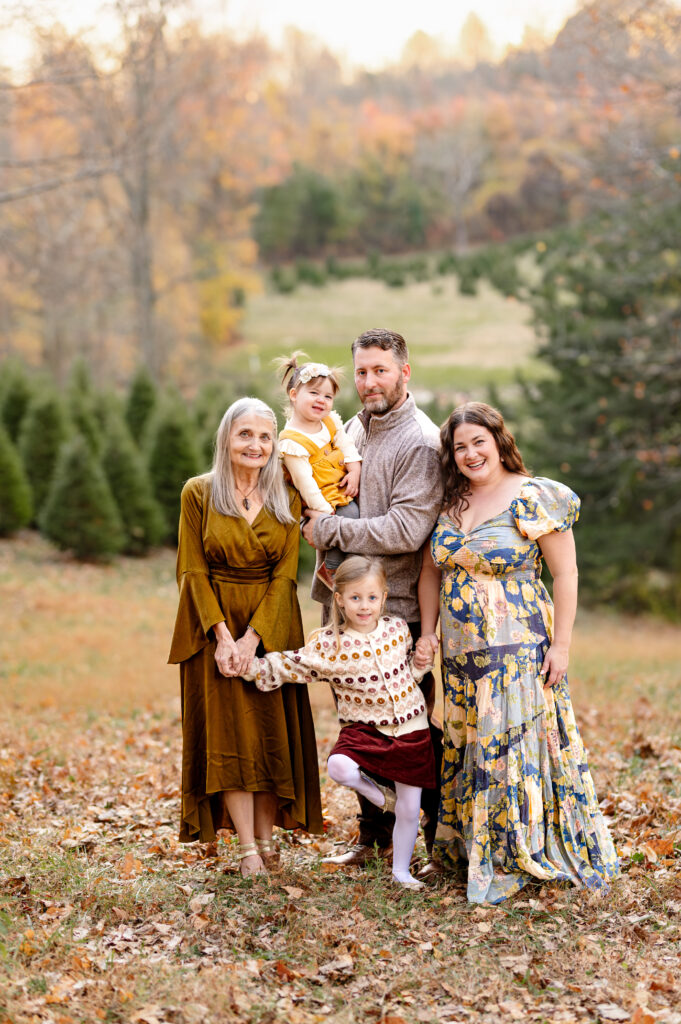 grandmother along with a family of 4 smiling together and standing with evergreen trees behind them