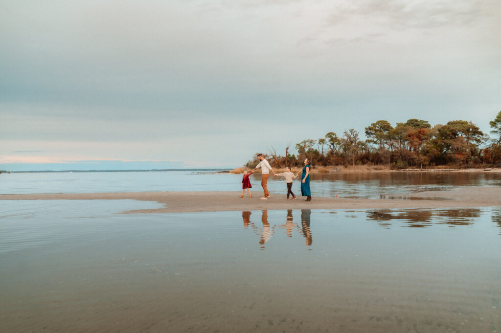 a family of 4 walks along a sliver of sand, with water surrounding them on all sides, their reflection is visible in the water