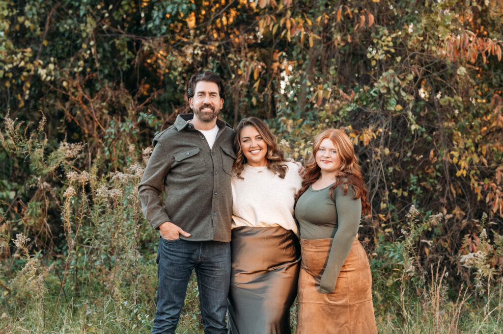 family of 3 stands in front of a tree smiling at the camera for a traditional portrait