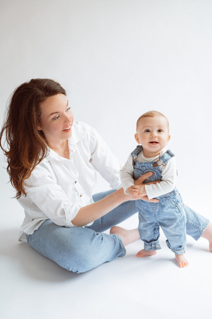 mom sitting on the floor against a white backdrop inside of a photo studio, she helps her infant daughter stand up