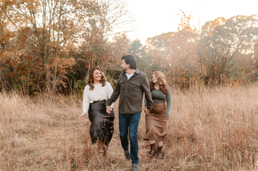 family of 3 walks and laughs with one another while they walk toward camera in the middle of a grassy field