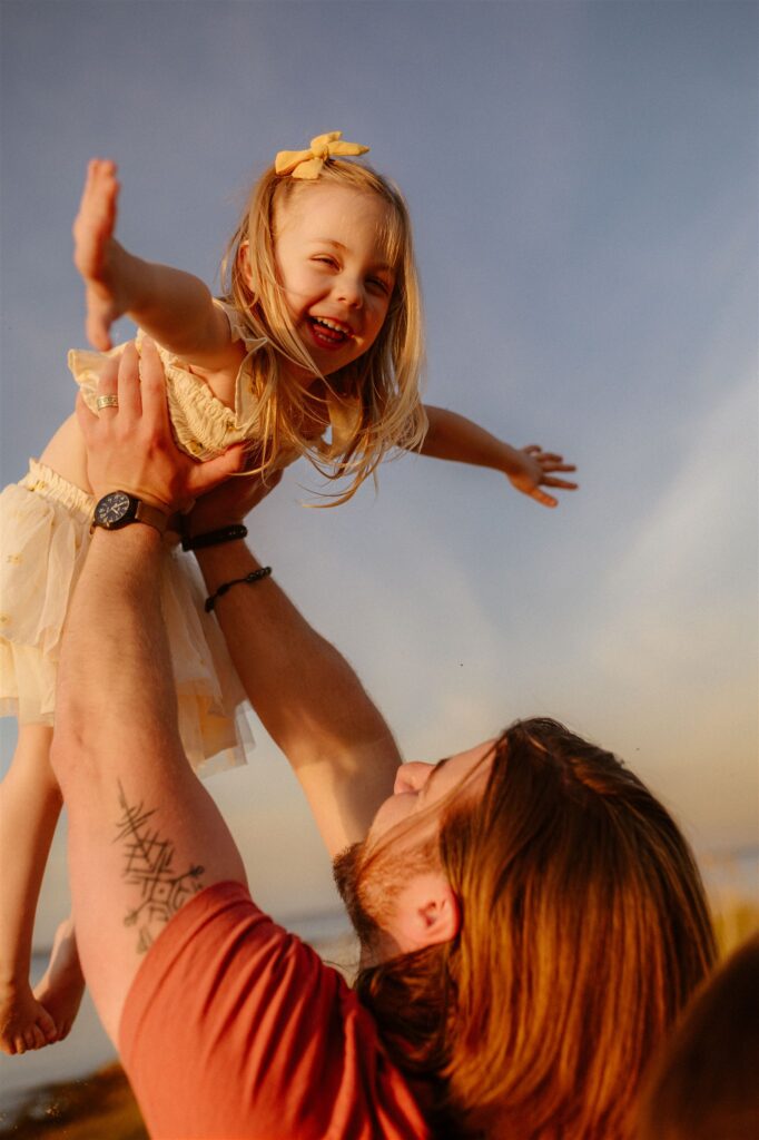Dad lifting laughing toddler girl high in the air during candid golden hour family photography session on Maryland beach