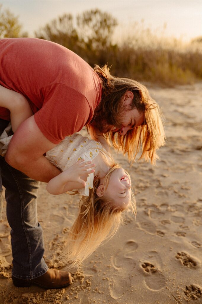 Dad flipping toddler daughter upside down on sandy beach during playful golden hour family session in Maryland