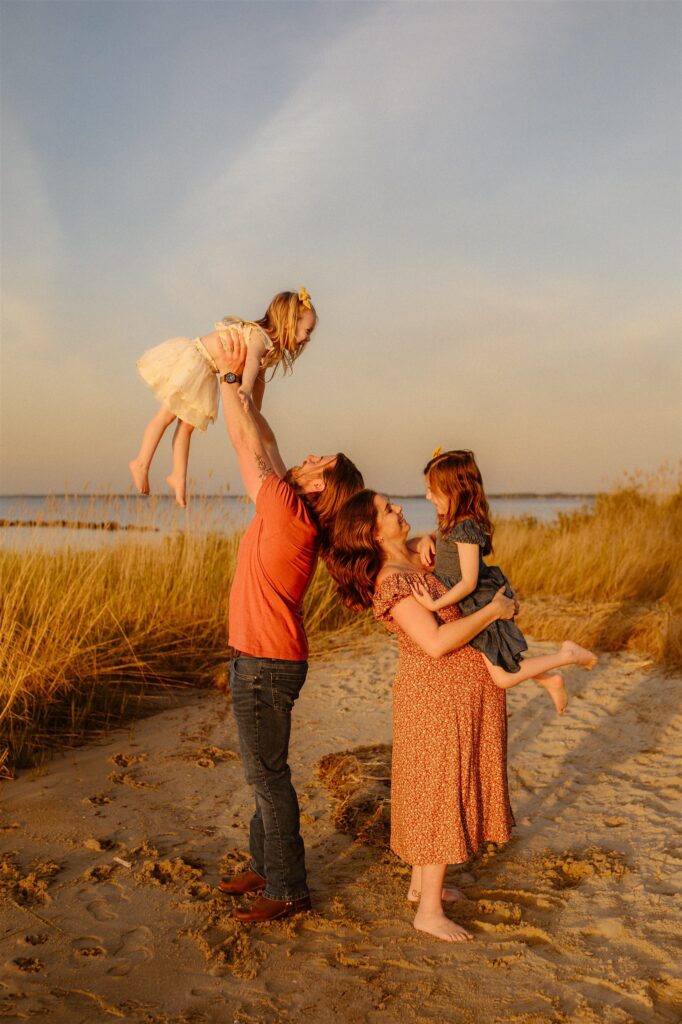 Family of four at golden hour on Maryland beach — dad lifting daughter in air while mom holds younger child in sand dunes