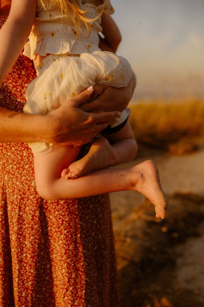 Mother holding toddler daughter with sandy feet during golden hour beach family session in Maryland